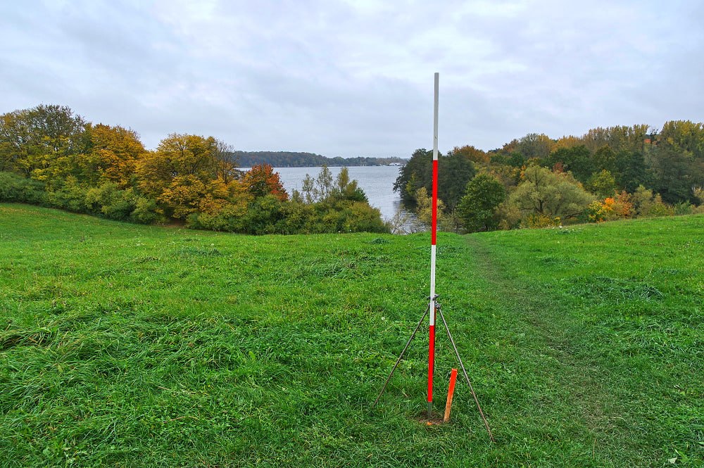 Roter und weißer Vermessungsstab auf einer Wiese mit Blick auf den Ziegelaußensee